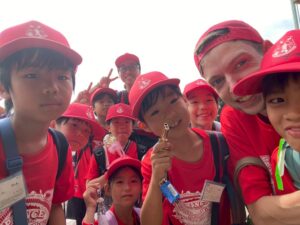 Group of children in red caps and shirts posing for a selfie with an adult supervisor outdoors, smiling and making peace signs.