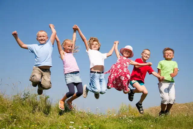 Six children leap in the air holding hands in a sunny grassy field, smiling under a blue sky, showing teamwork and fun.
